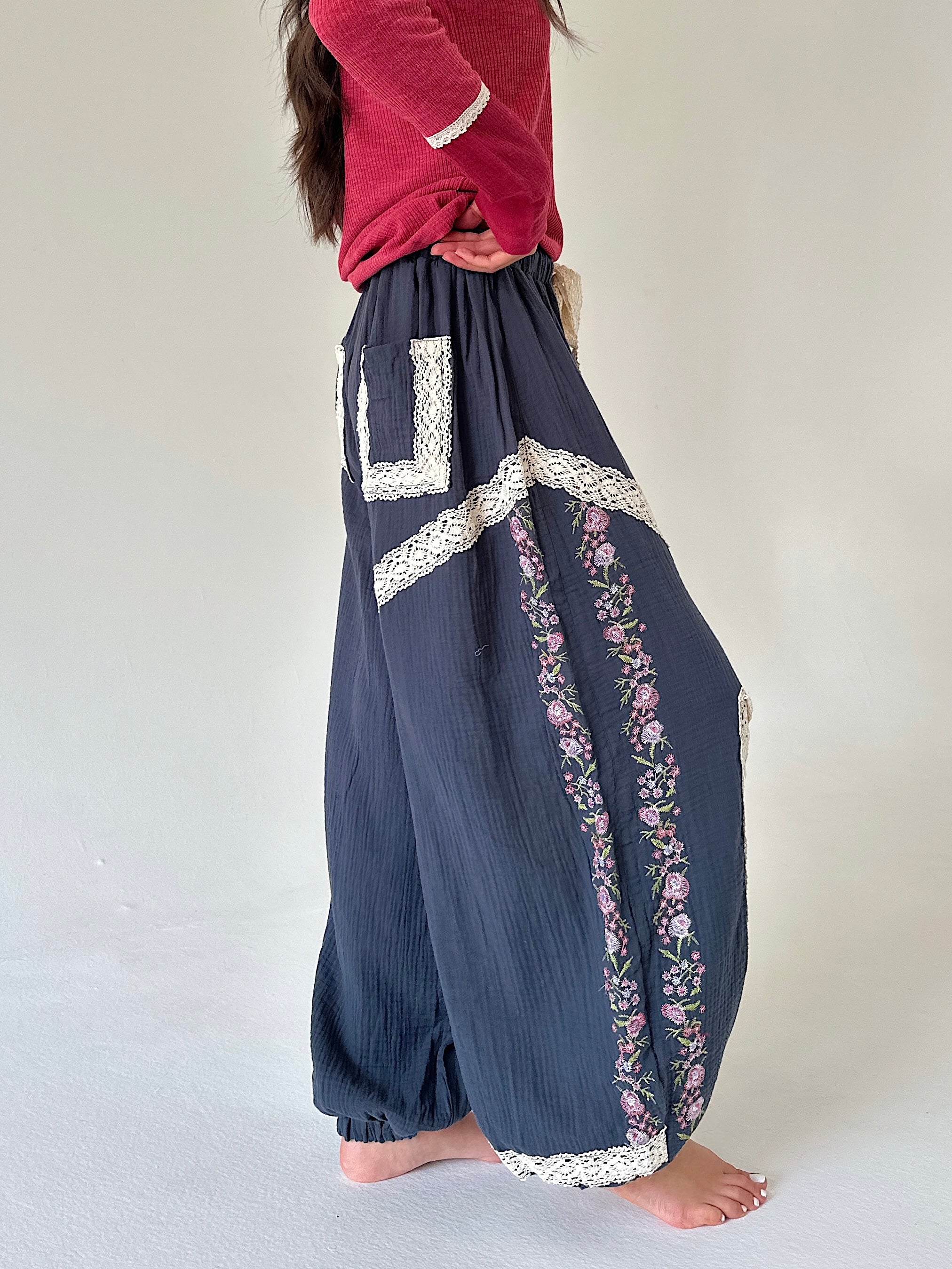 A person wearing loose navy blue harem-style pants with white lace trim and pink floral embroidery, paired with a red top, standing barefoot against a plain light background.