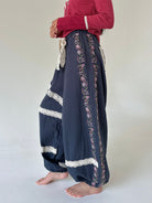 A person wearing loose, wide-leg navy pants with floral embroidery and white lace trim, paired with a red long-sleeve top, standing barefoot against a plain light background.