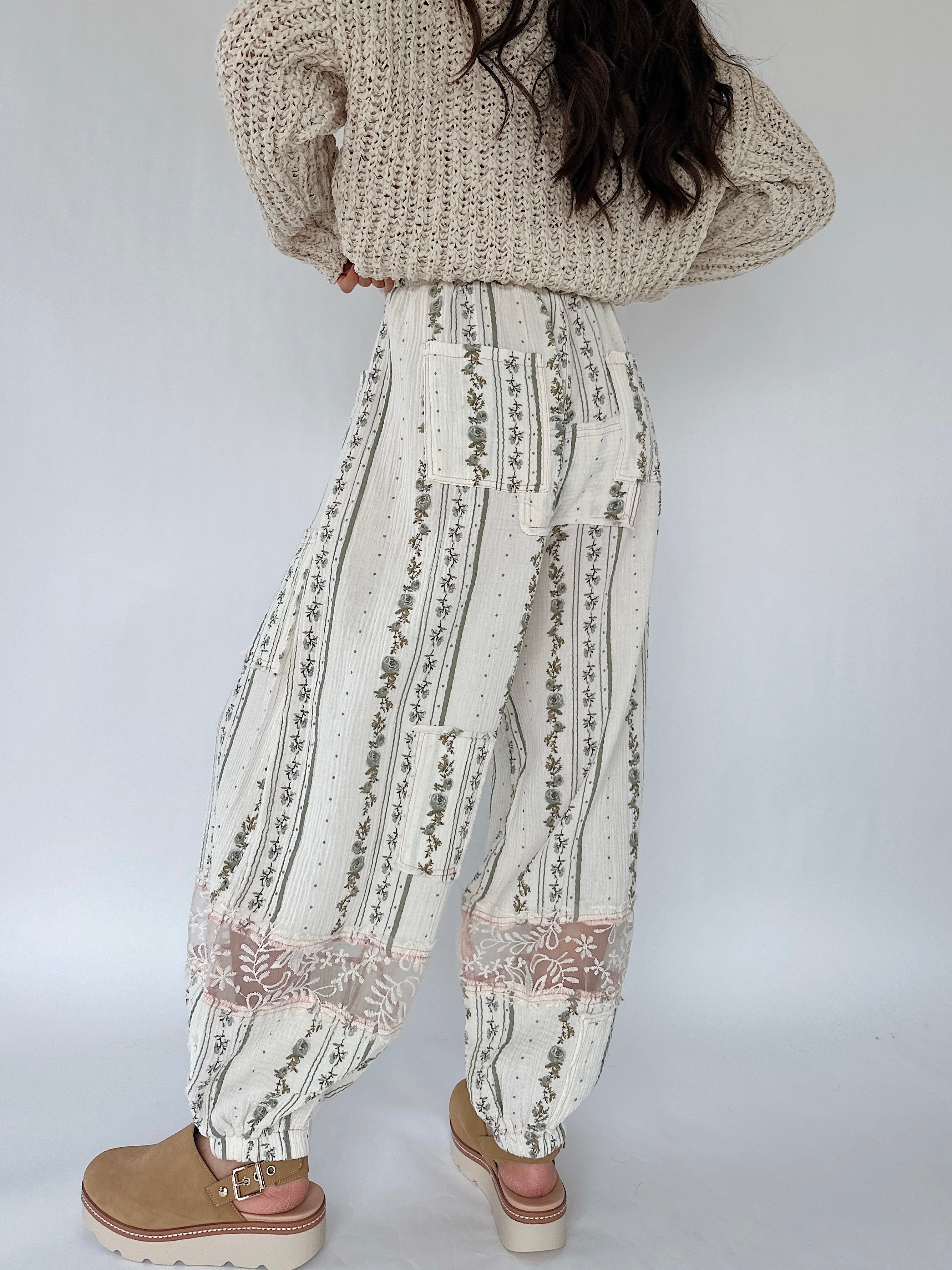 Person wearing a beige knit sweater and patterned pants on a white background from back