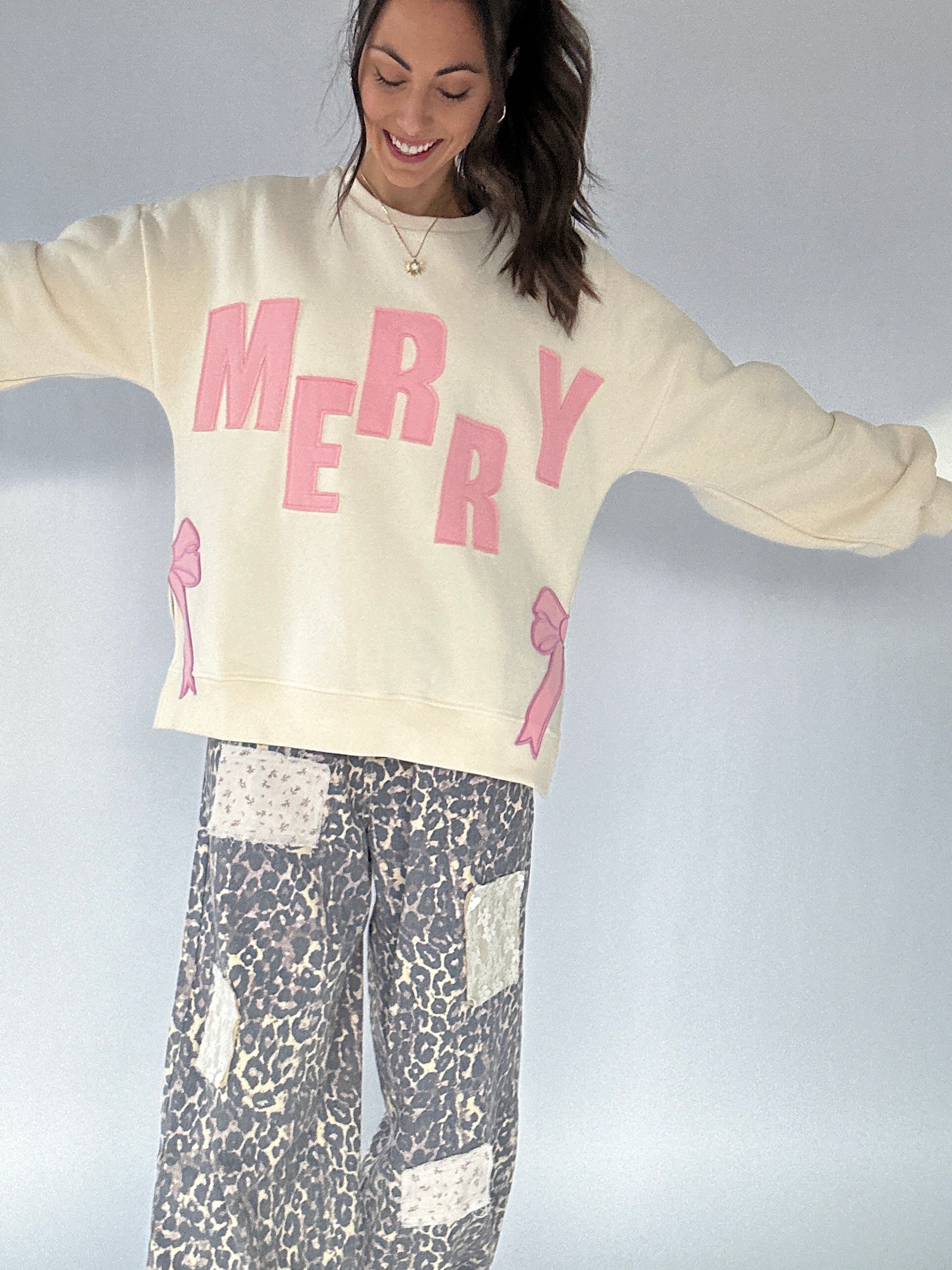 Person wearing a cream sweatshirt with 'MERRY' in pink and leopard patterned pants on a plain background