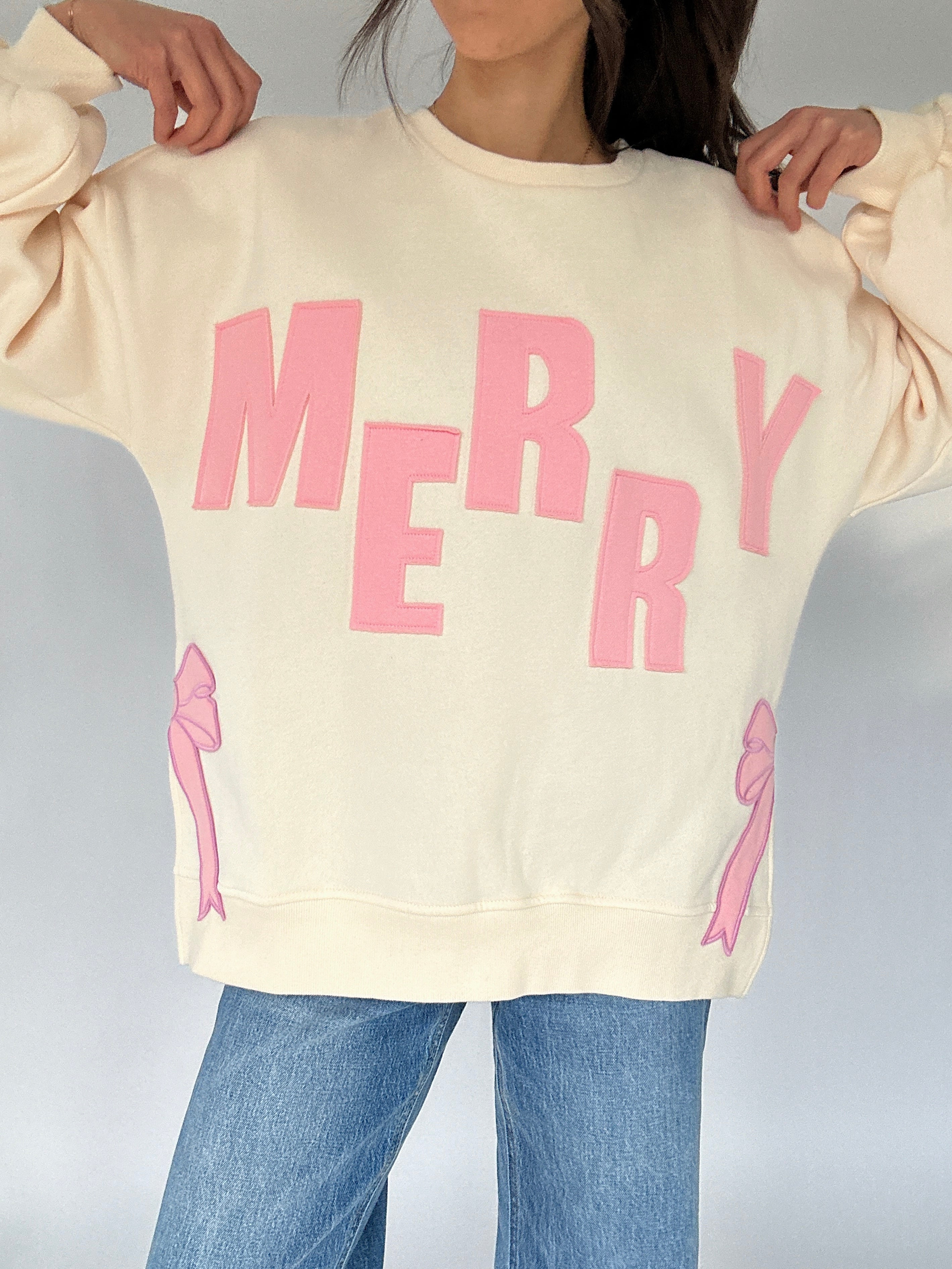 Beige sweatshirt with pink 'MERRY' text and bows on a plain background