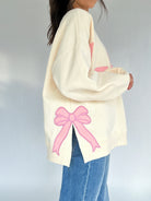 White sweatshirt with pink bow patch on side worn by a person on a plain background