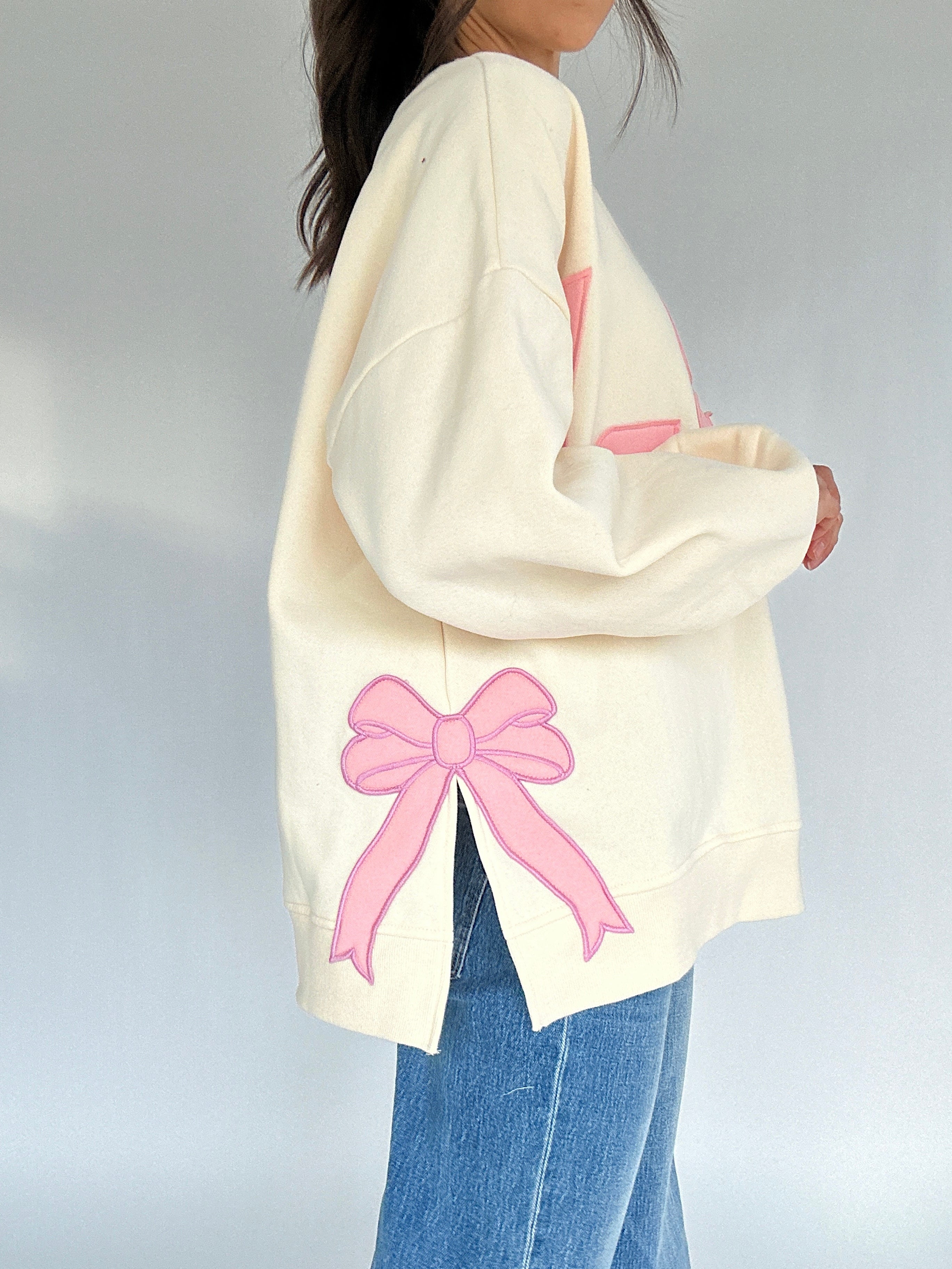 White sweatshirt with pink bow patch on side worn by a person on a plain background