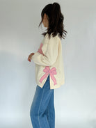 Person wearing a cream sweater with a pink bow patch on side and blue jeans on a white background