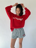 Person wearing a red sweater with 'Merry' text and checkered shorts on a white background putting hair in ponytail
