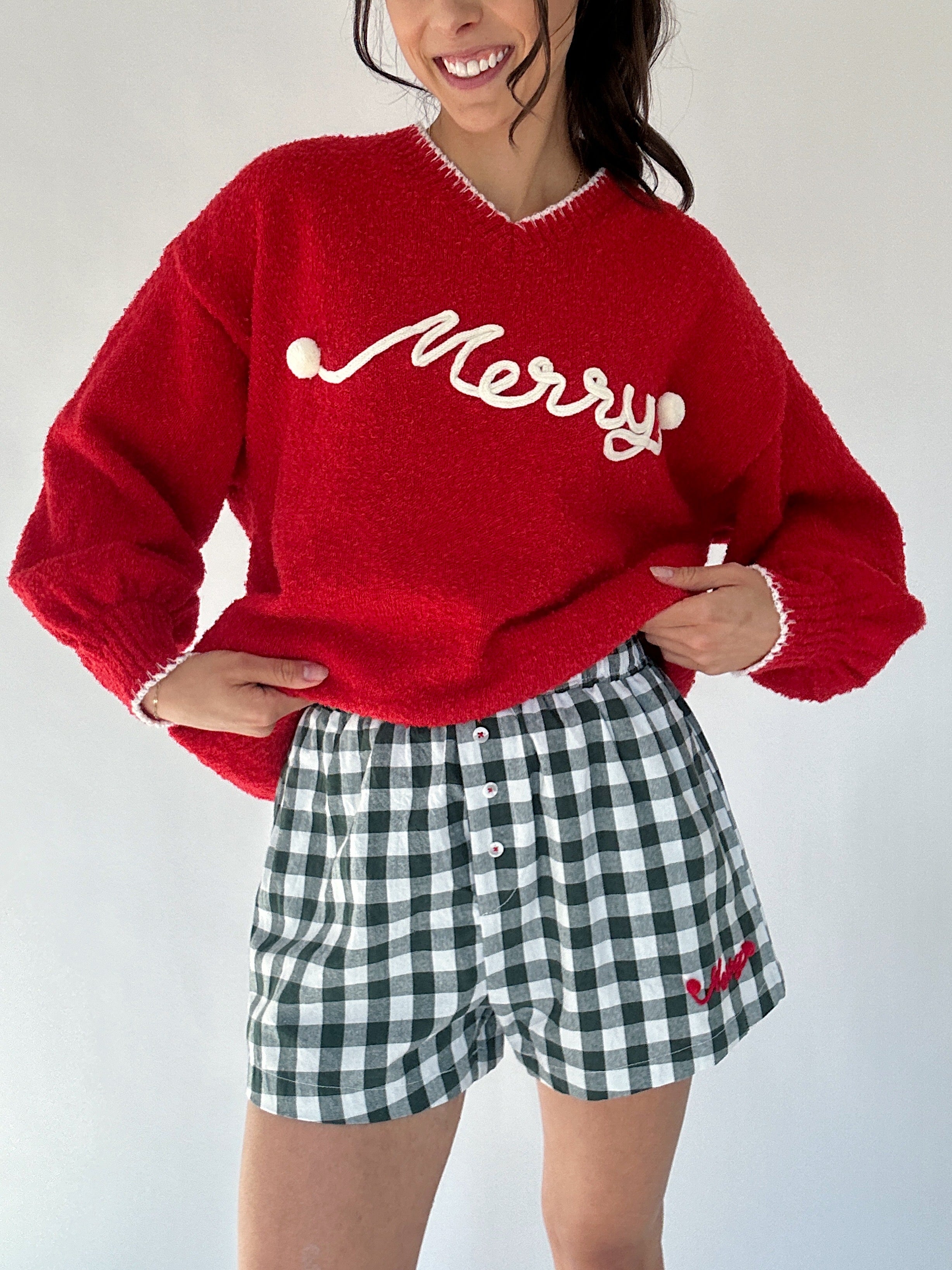 Person wearing a red sweater with 'Merry' stitched lettering and green checkered shorts on a white background
