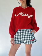 Person wearing a red sweater with white 'Merry' text and green checkered shorts on a white background