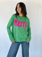 A woman is wearing a green knit sweater with the word 'Merry' in hot pink lettering across the front. She is also wearing blue jeans.