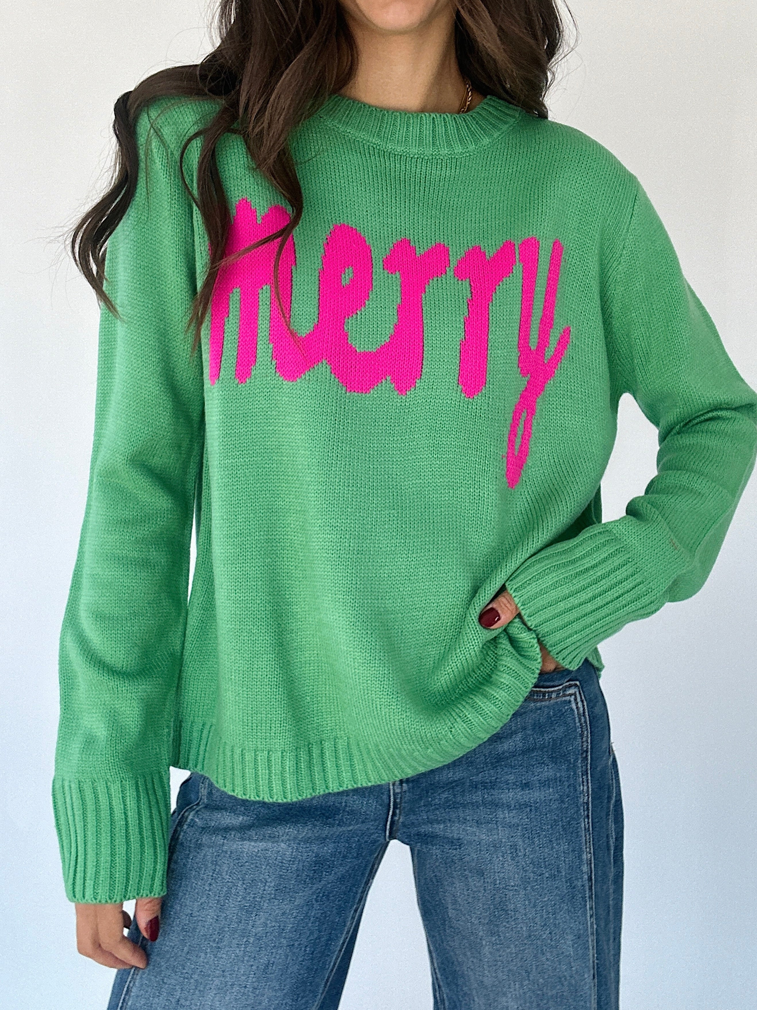 Green sweater with pink "merry" lettering with blue jeans on a plain background