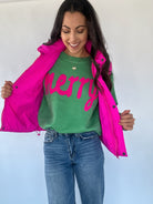 Woman wearing a green sweater with pink 'merry' lettering and a pink puffer vest on a plain background