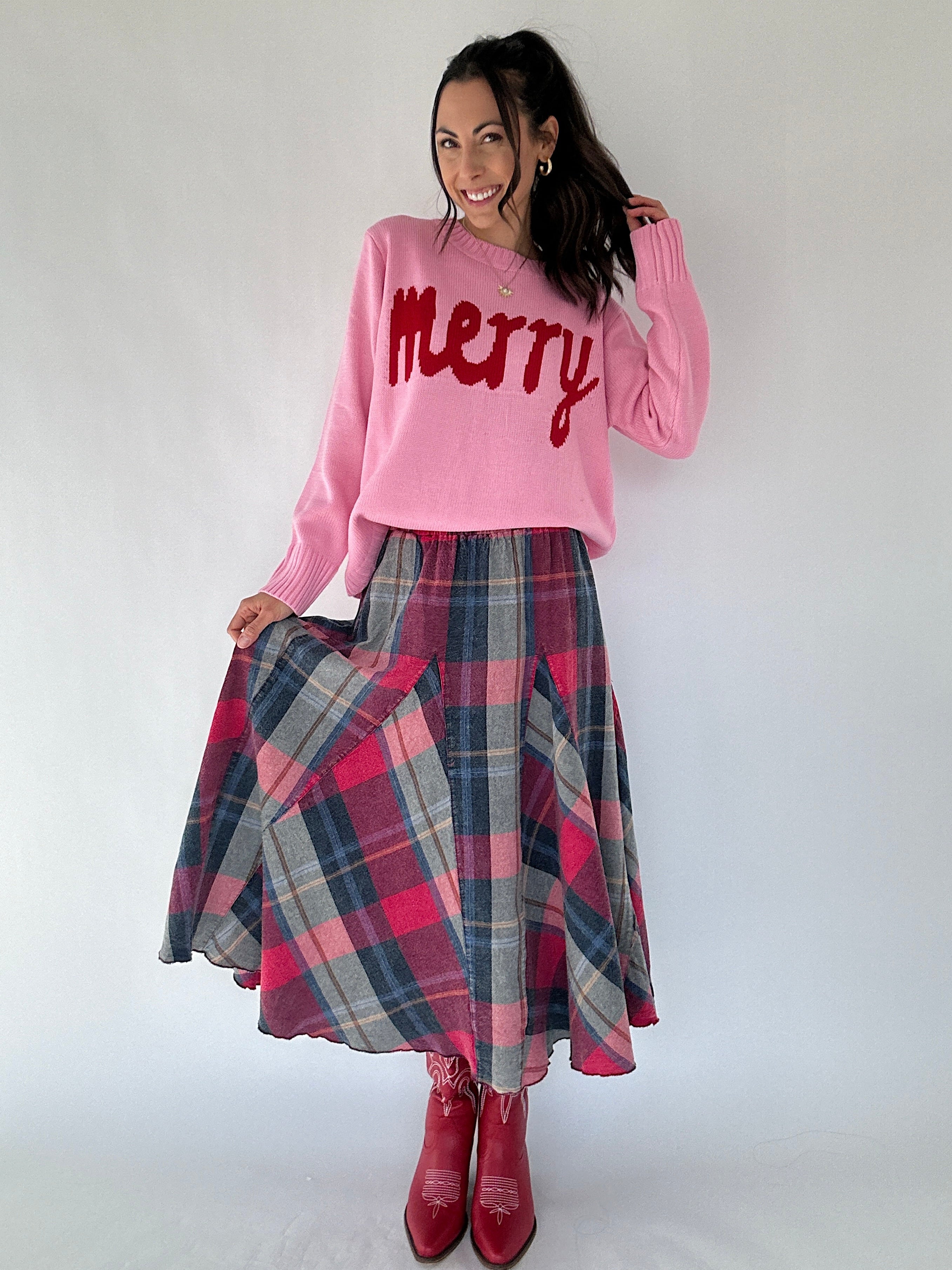 Woman wearing a pink sweater with 'Merry' text and a plaid skirt on a white background