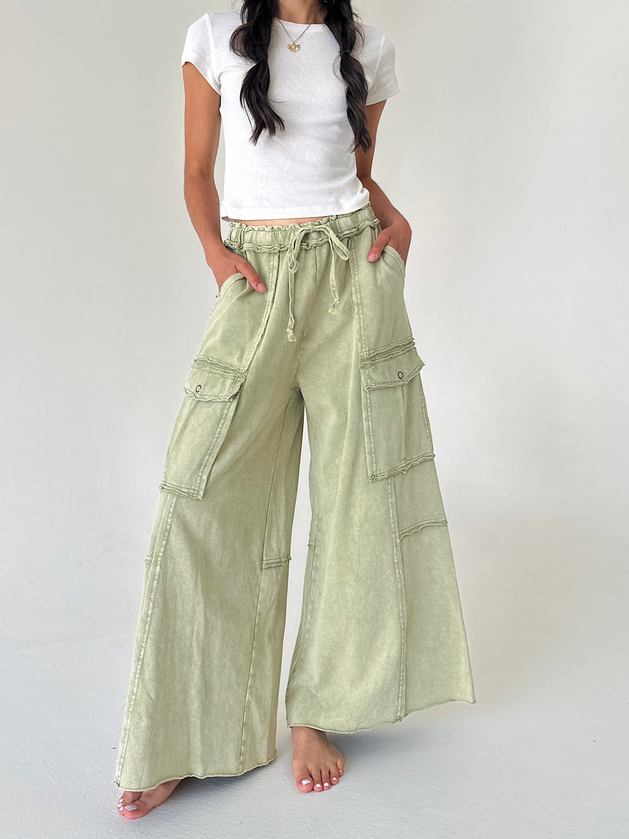 A model wearing light sage green wide-leg cargo pants with a drawstring waist, paired with a white t-shirt, standing against a plain light gray studio background.