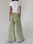 A person shown from the back wearing wide-leg sage green pants with a white t-shirt, standing against a plain light gray studio background.