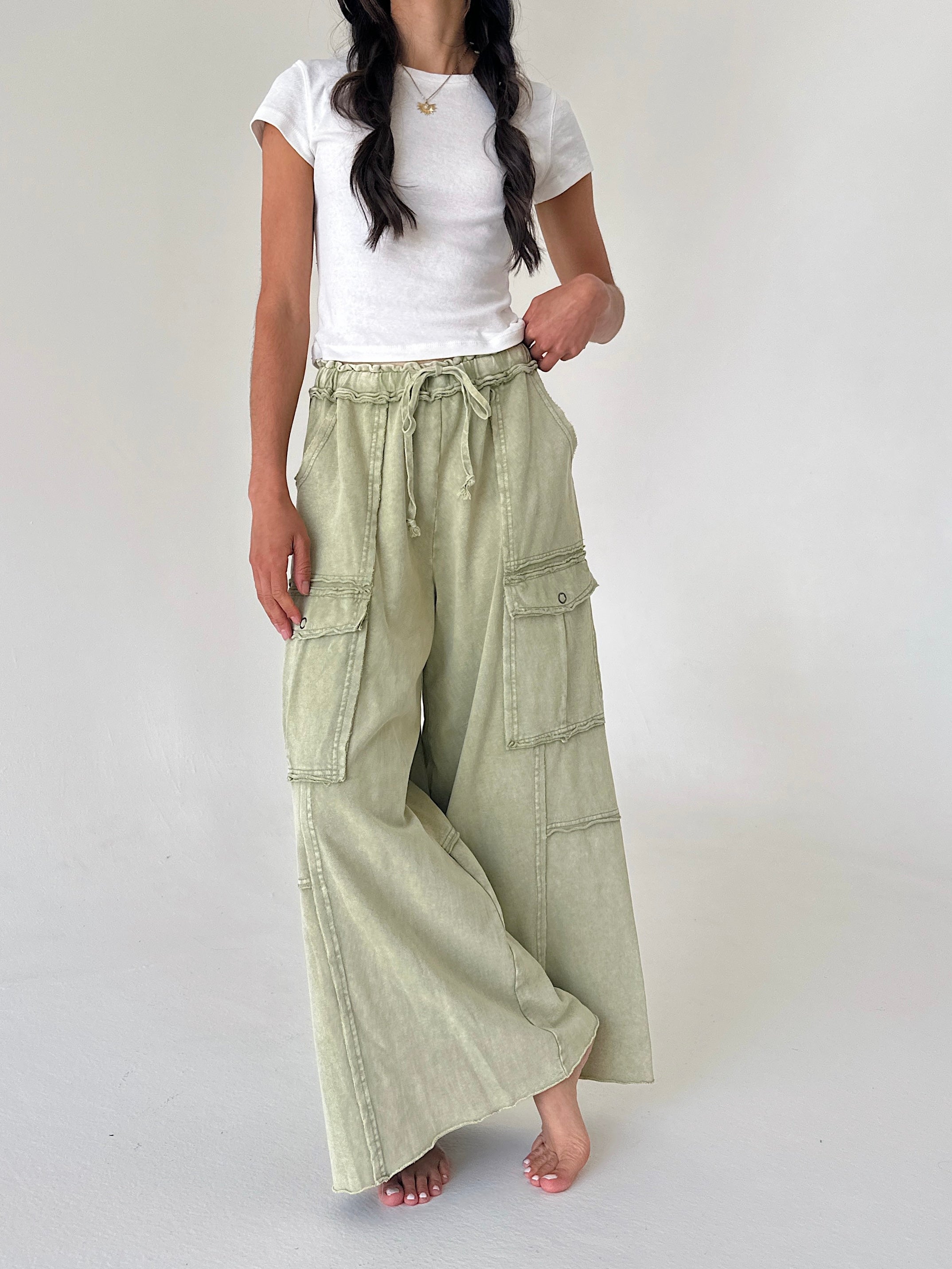 A woman wearing wide-leg sage green cargo pants with a drawstring waist, styled with a white t-shirt, standing against a plain light gray studio background.