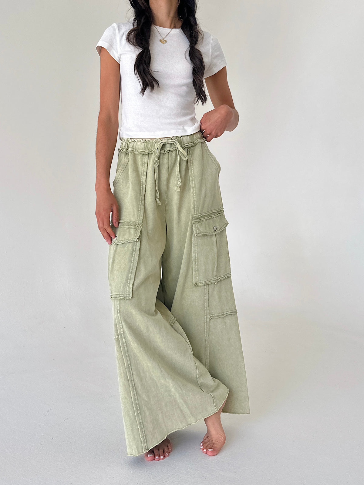 A woman wearing wide-leg sage green cargo pants with a drawstring waist, styled with a white t-shirt, standing against a plain light gray studio background.
