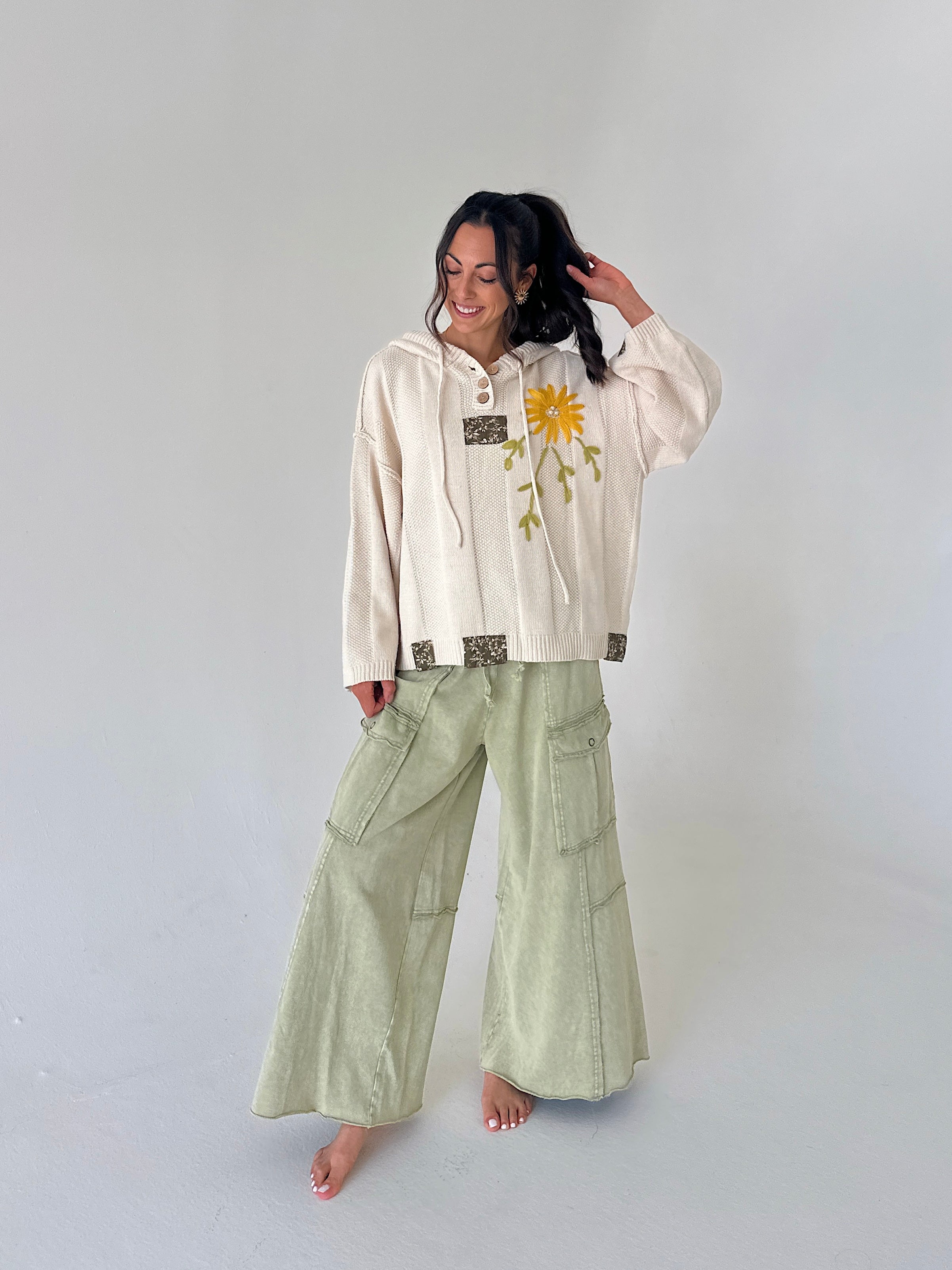 A woman stands barefoot against a light gray studio background, wearing a cream hooded sweatshirt with a yellow flower graphic and green wide-leg cargo pants.