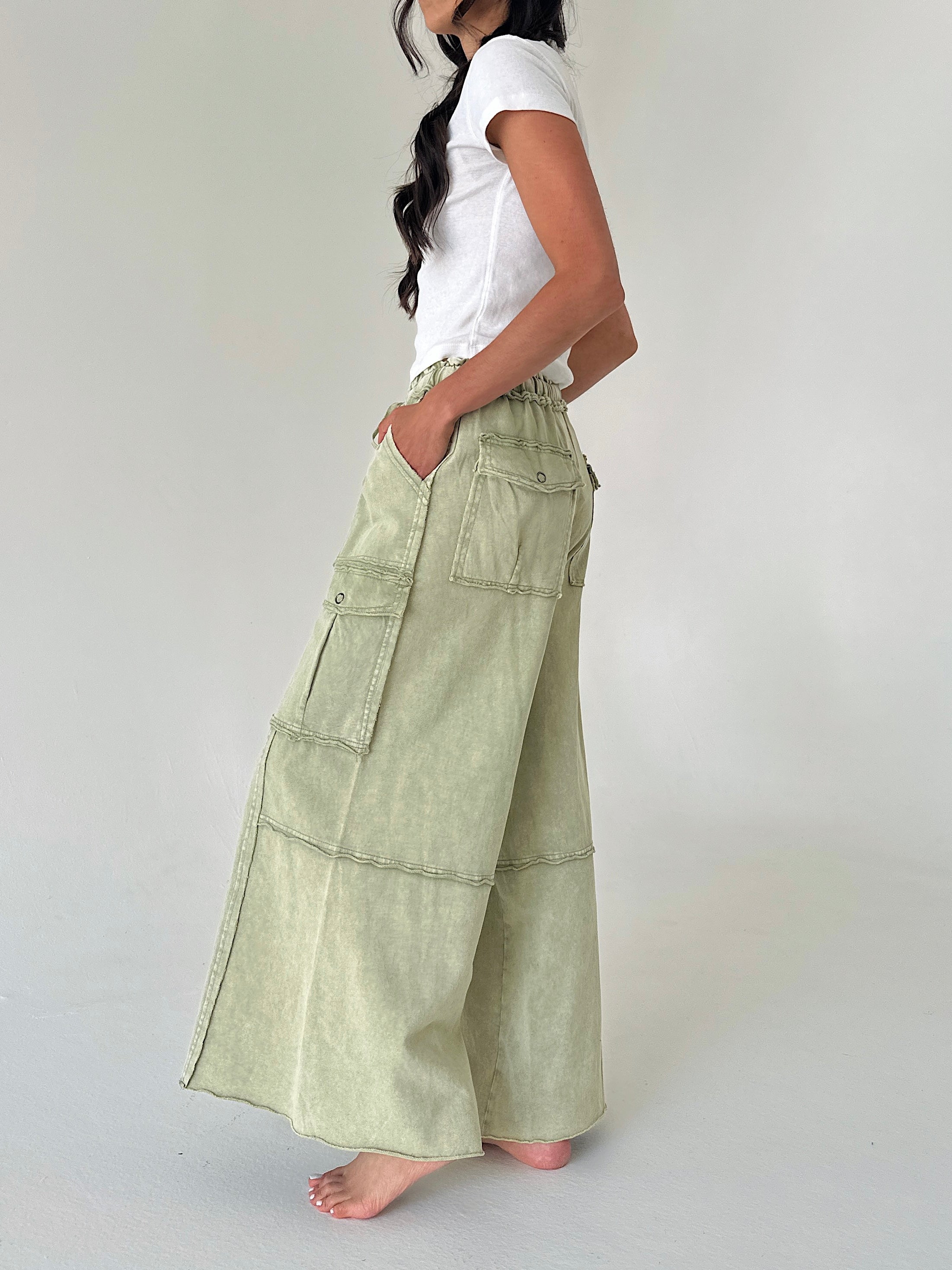 A woman wearing a white t-shirt and light sage green wide-leg cargo pants, standing barefoot against a plain light gray studio background.