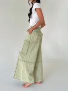 A woman wearing a white t-shirt and light sage green wide-leg cargo pants, standing barefoot against a plain light gray studio background.