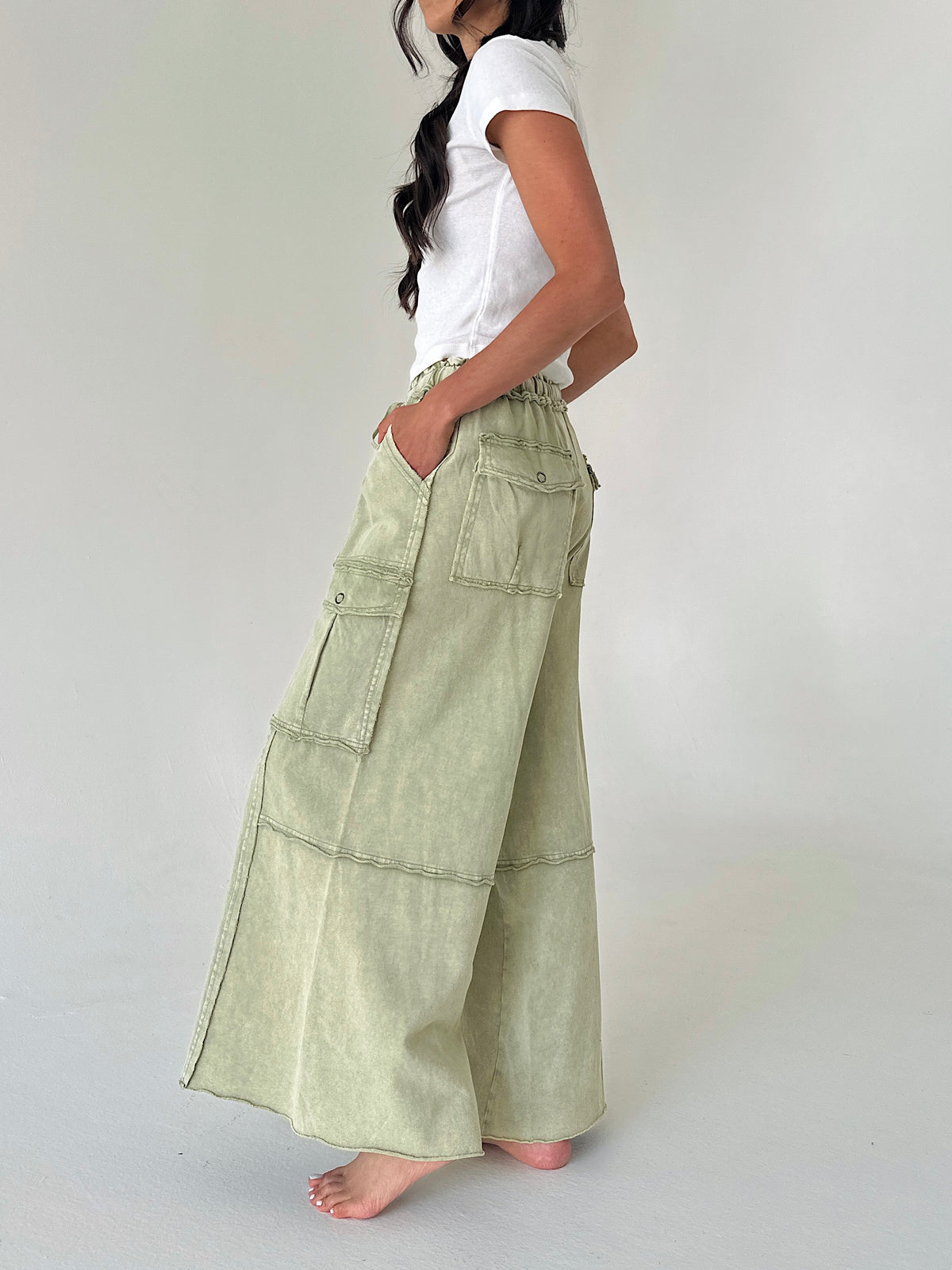 A woman wearing a white t-shirt and light sage green wide-leg cargo pants, standing barefoot against a plain light gray studio background.