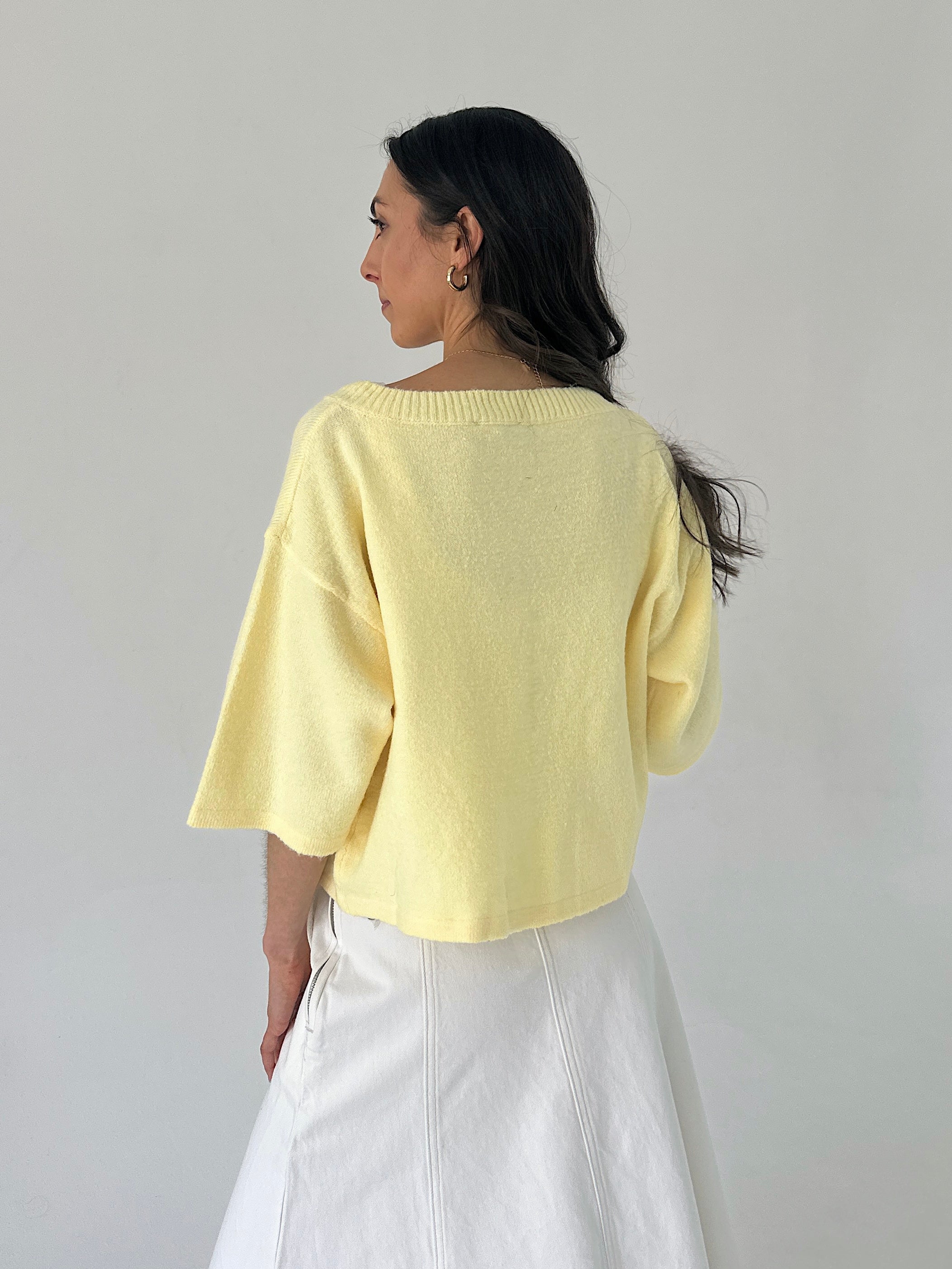 Woman wearing a light yellow sweater and white pants against a plain background showing back