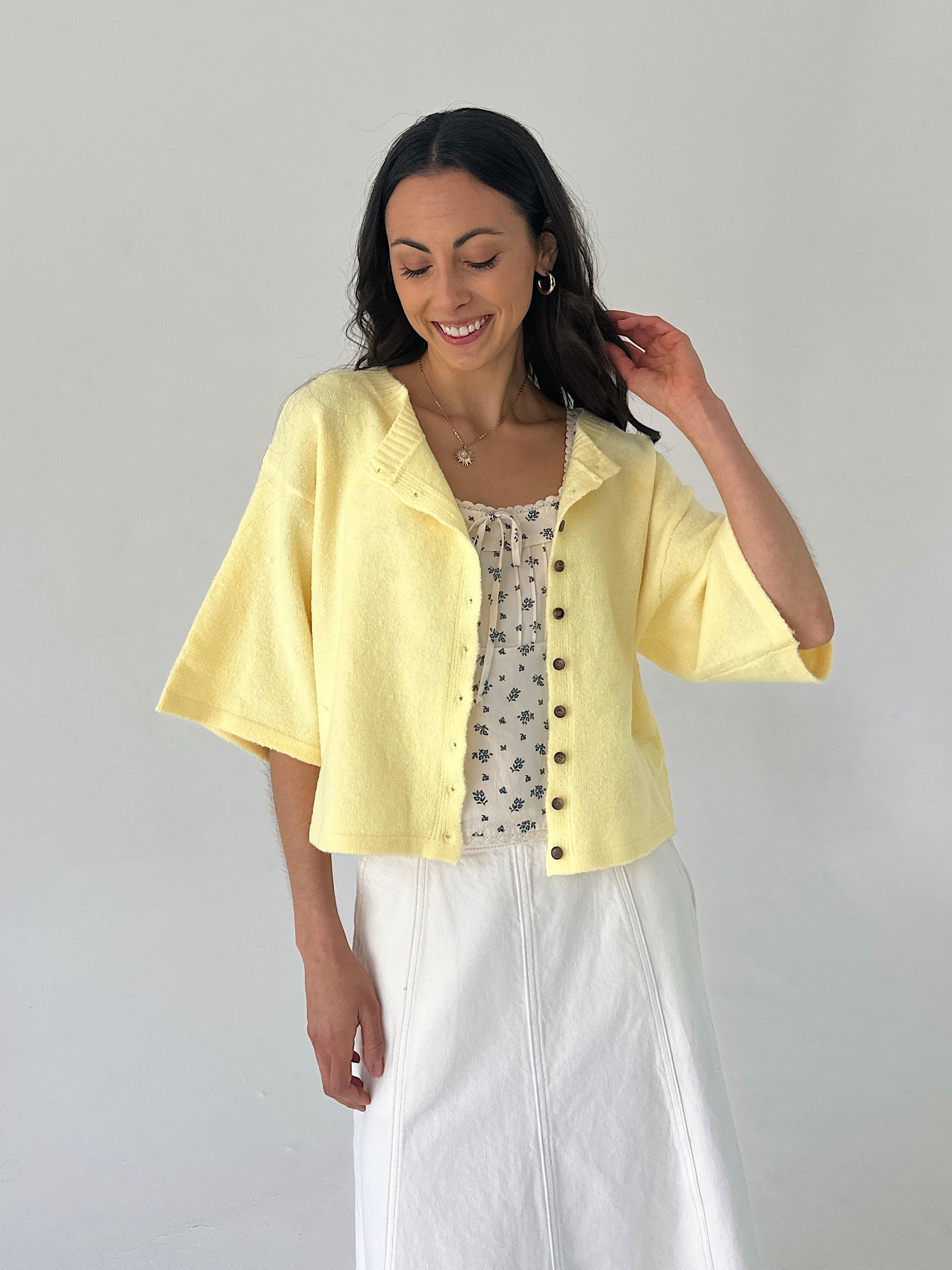 Woman wearing a light yellow cardigan over a patterned top and white skirt on a plain background
