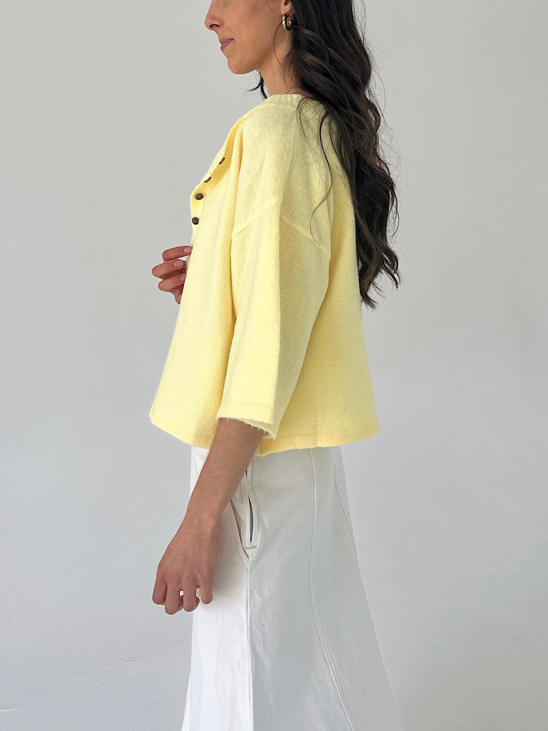 Woman wearing a light yellow cardigan and white pants against a plain background from side