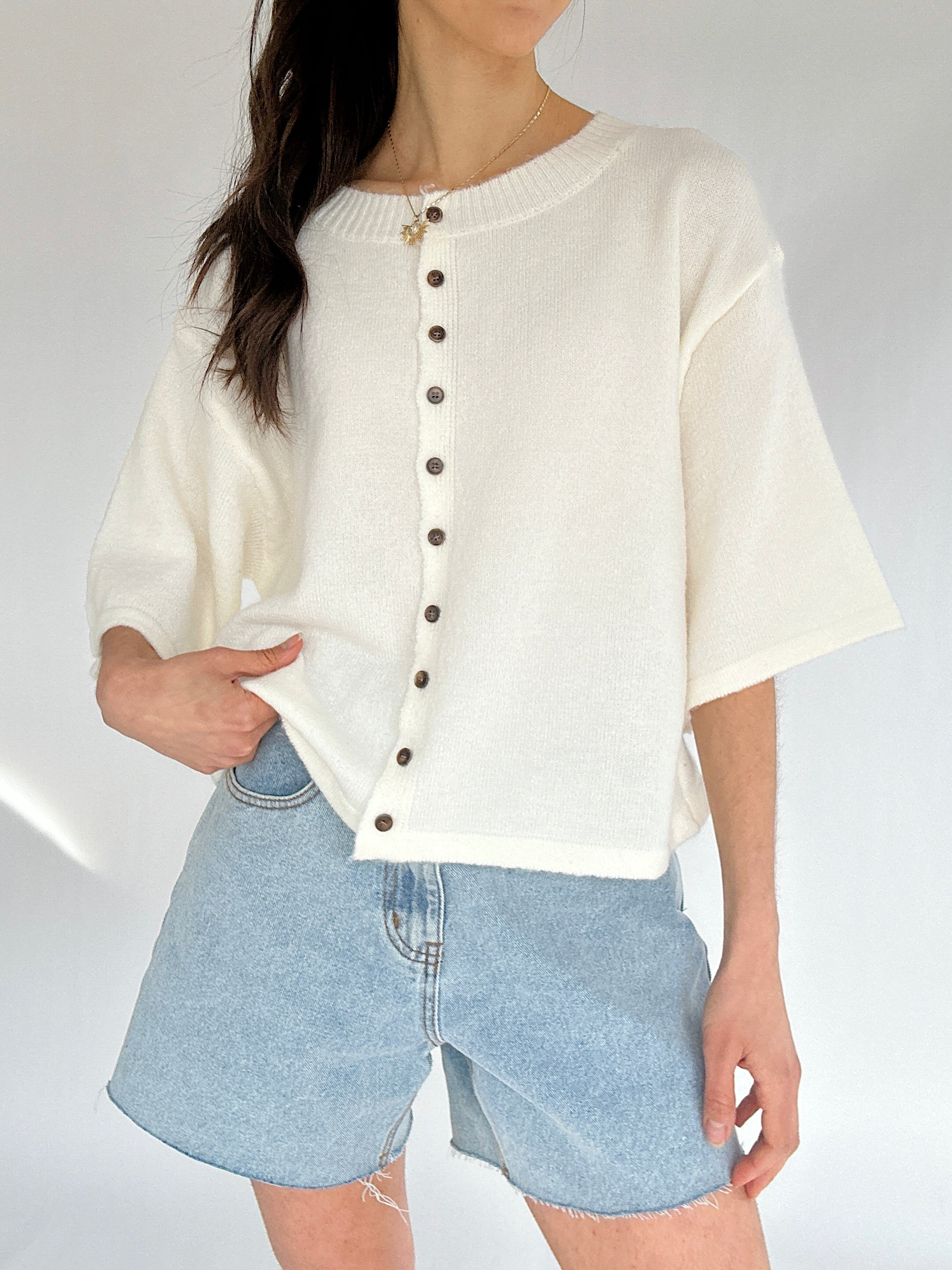 Person wearing a white button-up short sleeve cardigan and denim shorts on a plain background