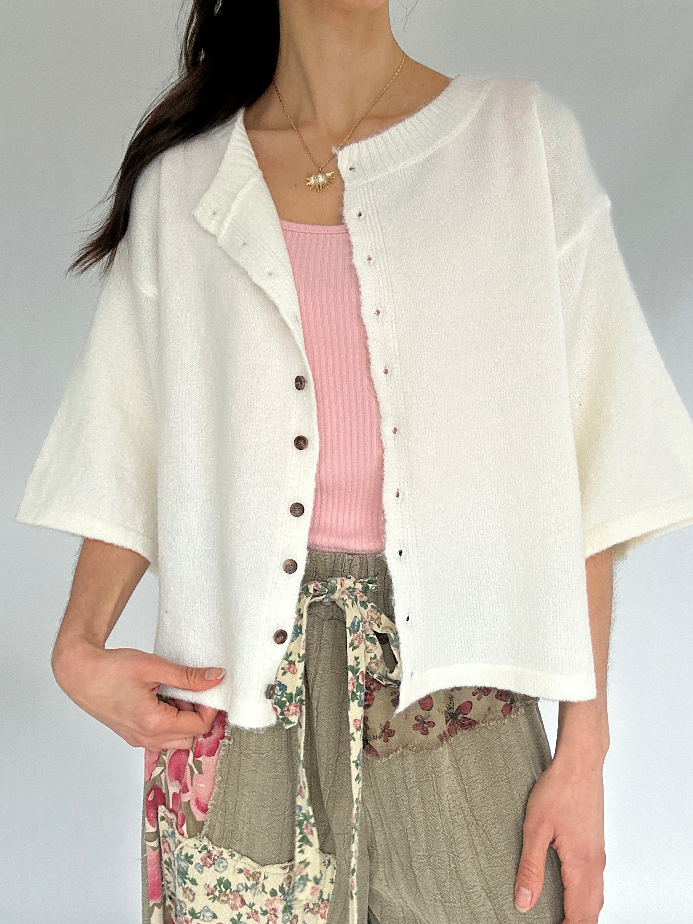 Person wearing a white cardigan over a pink top and floral pants on a gray background
