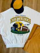 WV Mountaineers Football graphic on a white sweatshirt with a ballcap and pair of pants folded on the floor.