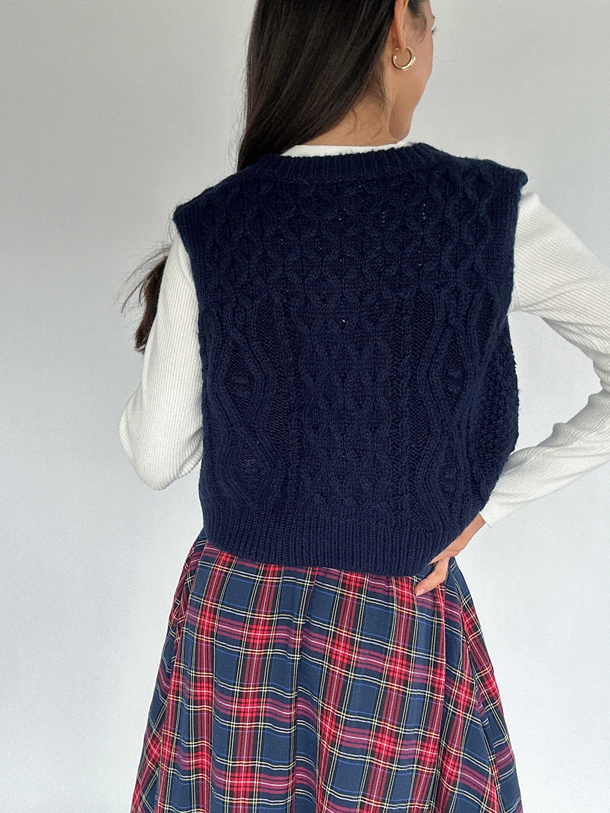 Person wearing a navy blue knitted vest over a white shirt and plaid skirt on a plain background showing solid back