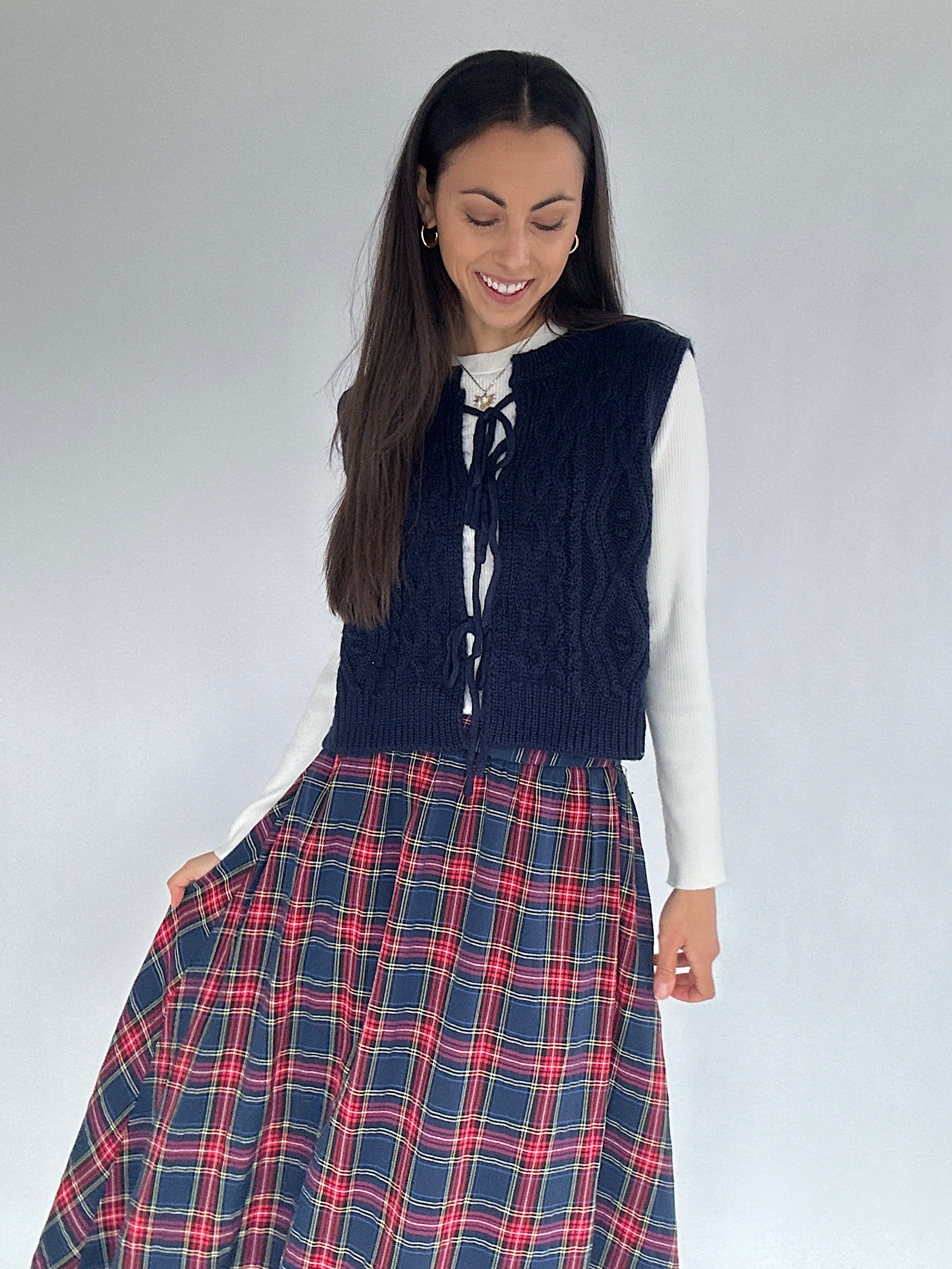 Woman wearing a navy cable knit vest with tie front over a white long sleeve and red and blue plaid skirt on a plain background