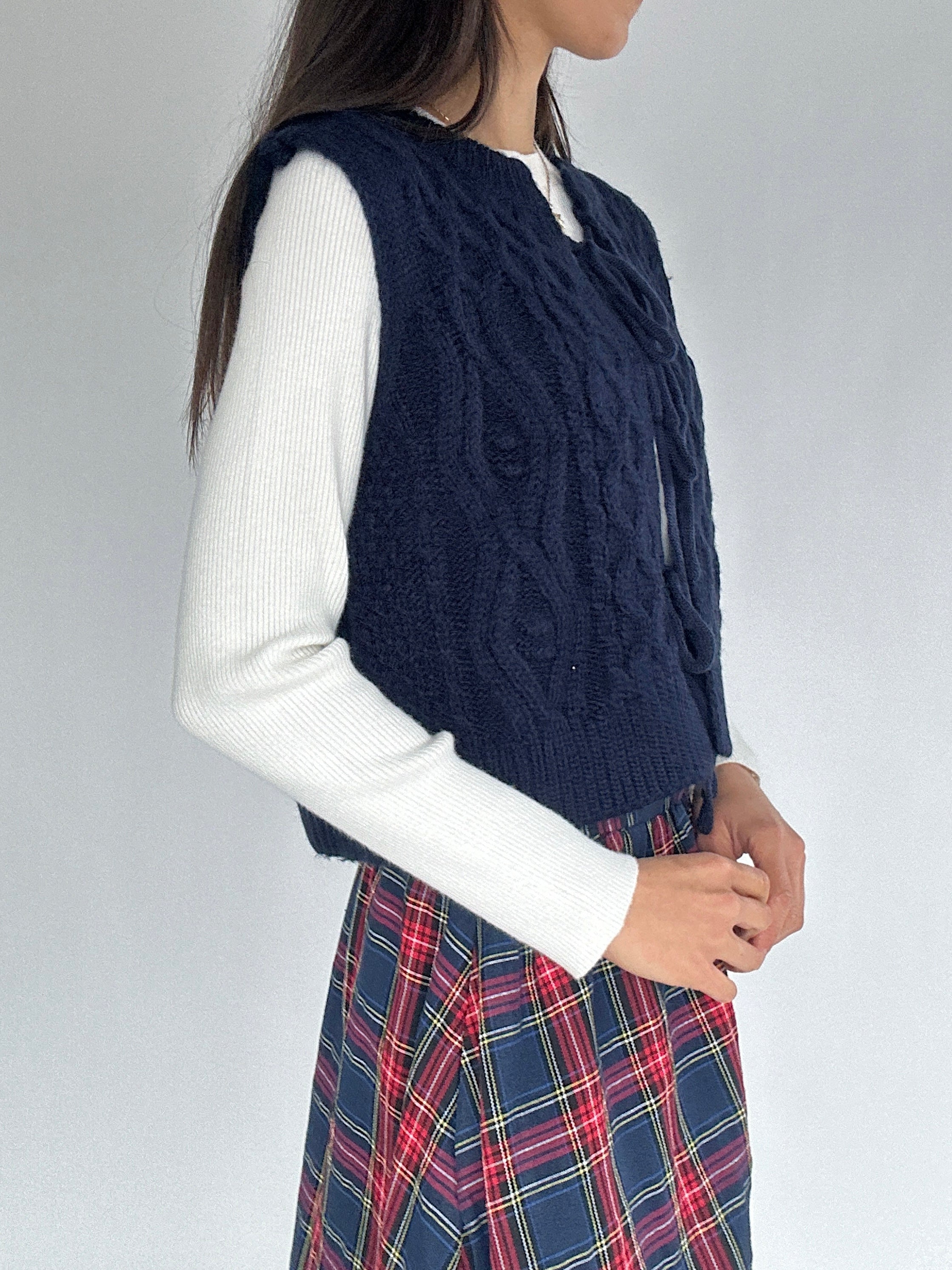 Person wearing a navy knitted vest over a white long-sleeve shirt and plaid skirt on a plain background showing side