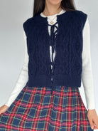 Person wearing a navy blue knitted vest over a white shirt and plaid skirt on a plain background