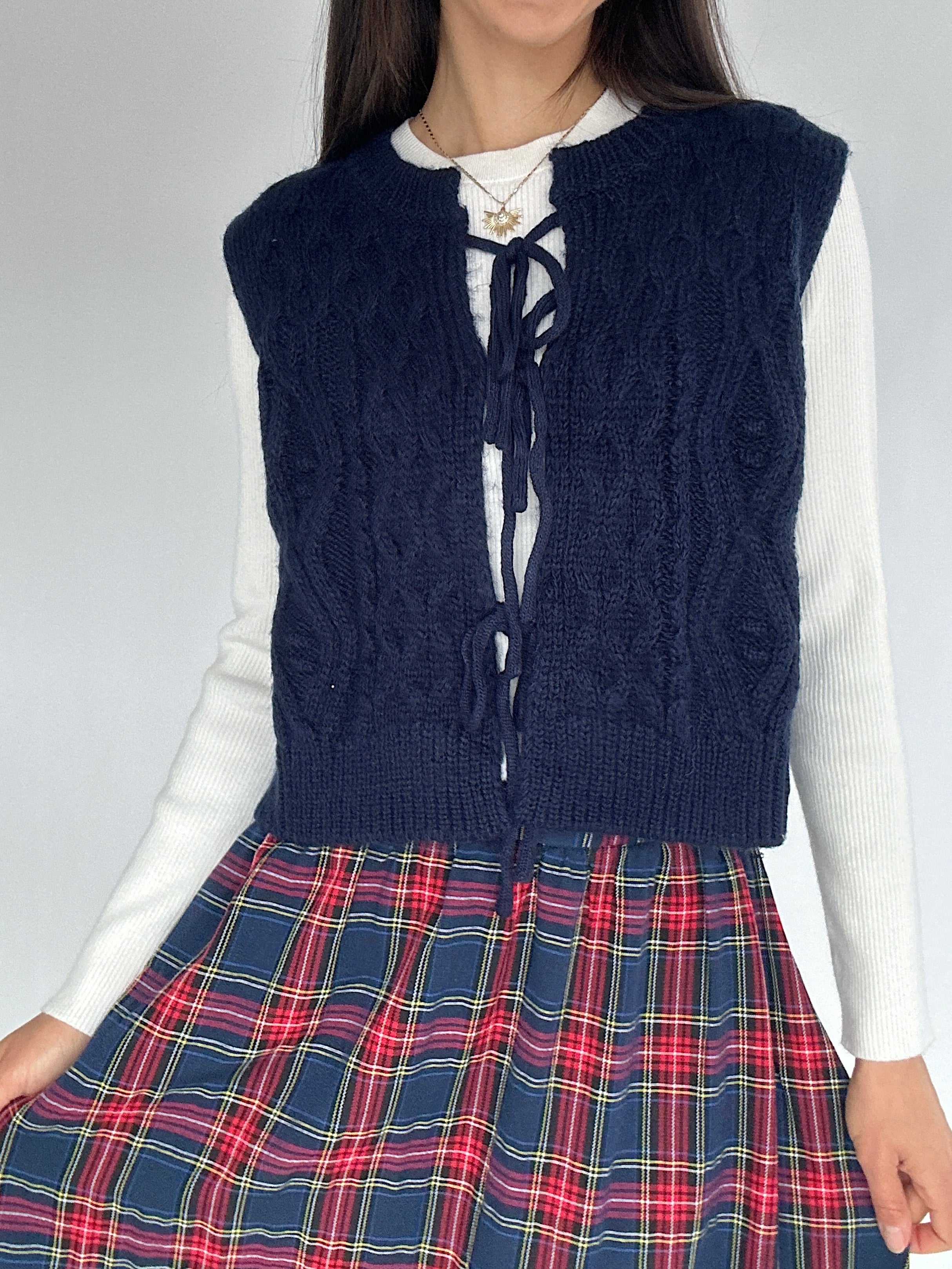 Person wearing a navy blue knitted vest over a white shirt and plaid skirt on a plain background