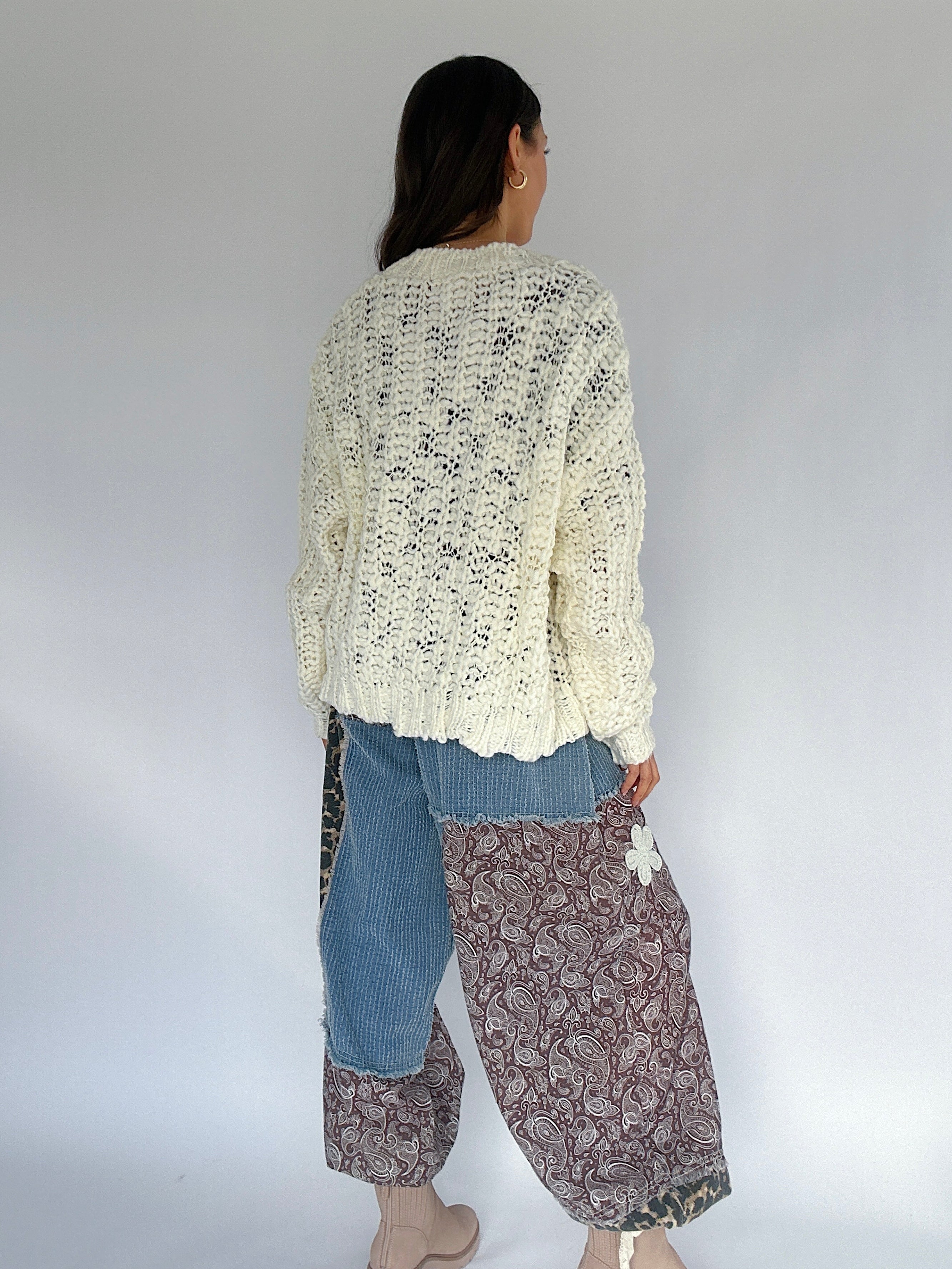 Person wearing a cream knitted cardigan, blue jeans, and patterned pants against a white background from back