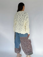 Person wearing a cream knitted cardigan, blue jeans, and patterned pants against a white background from back