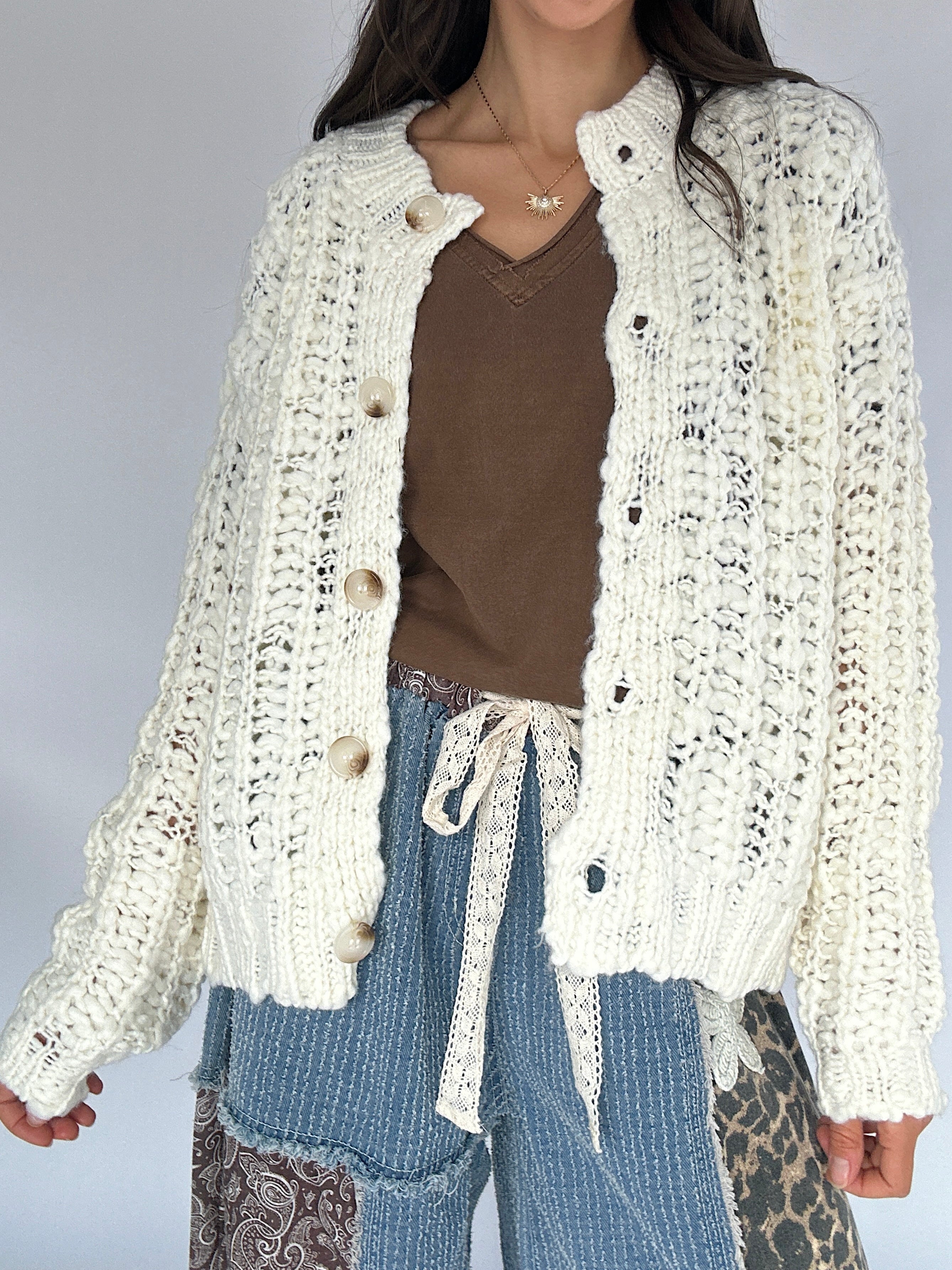 Person wearing a cream crocheted cardigan over a brown top and blue jeans on a gray background