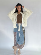 Woman wearing a cream cardigan, brown top, blue jeans, and patterned pants on a white background