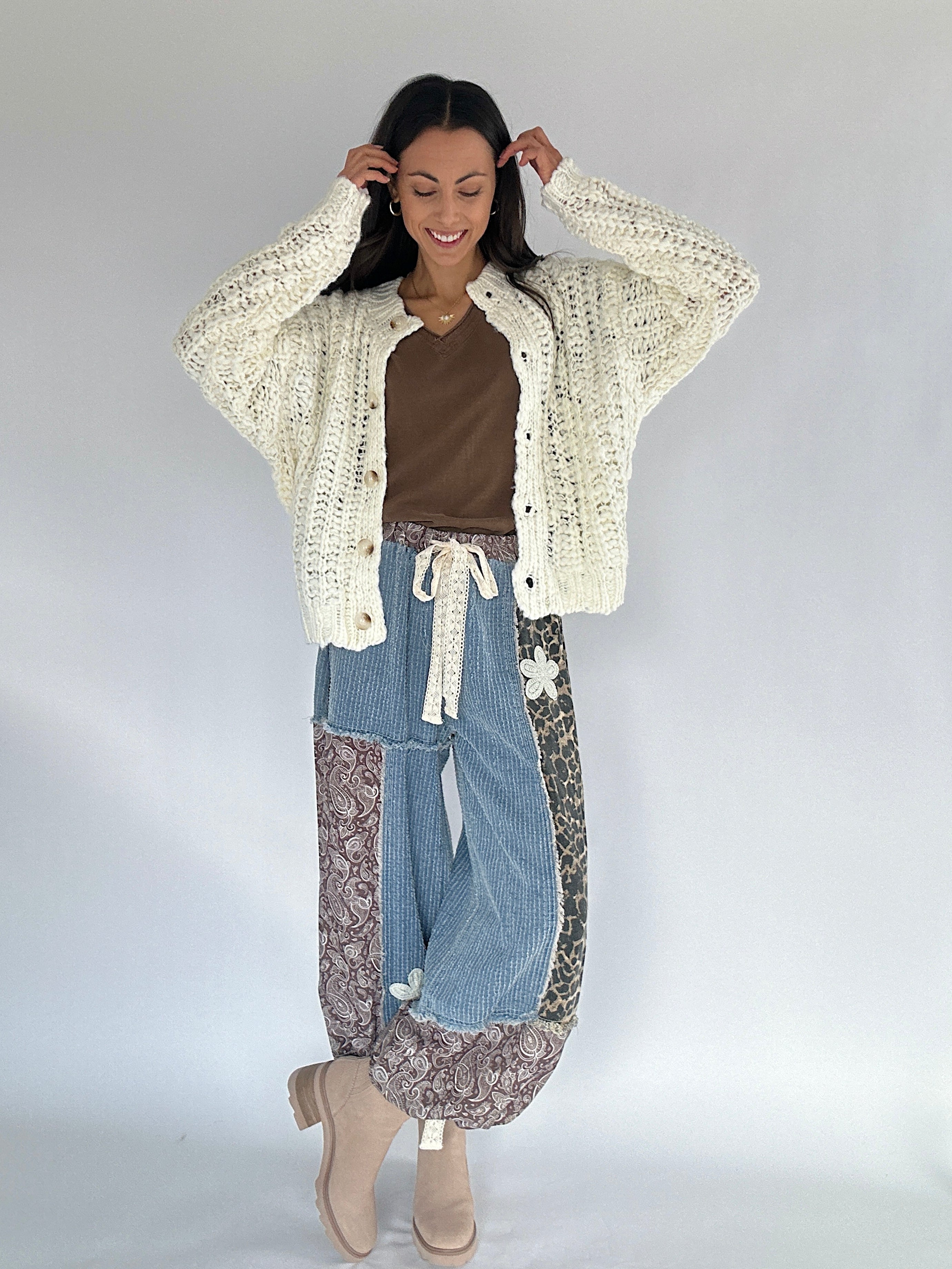 Woman wearing a cream cardigan, brown top, blue jeans, and patterned pants on a white background