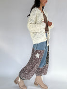 Woman wearing a cream crochet cardigan over a brown top with patterned pants on a white background from side