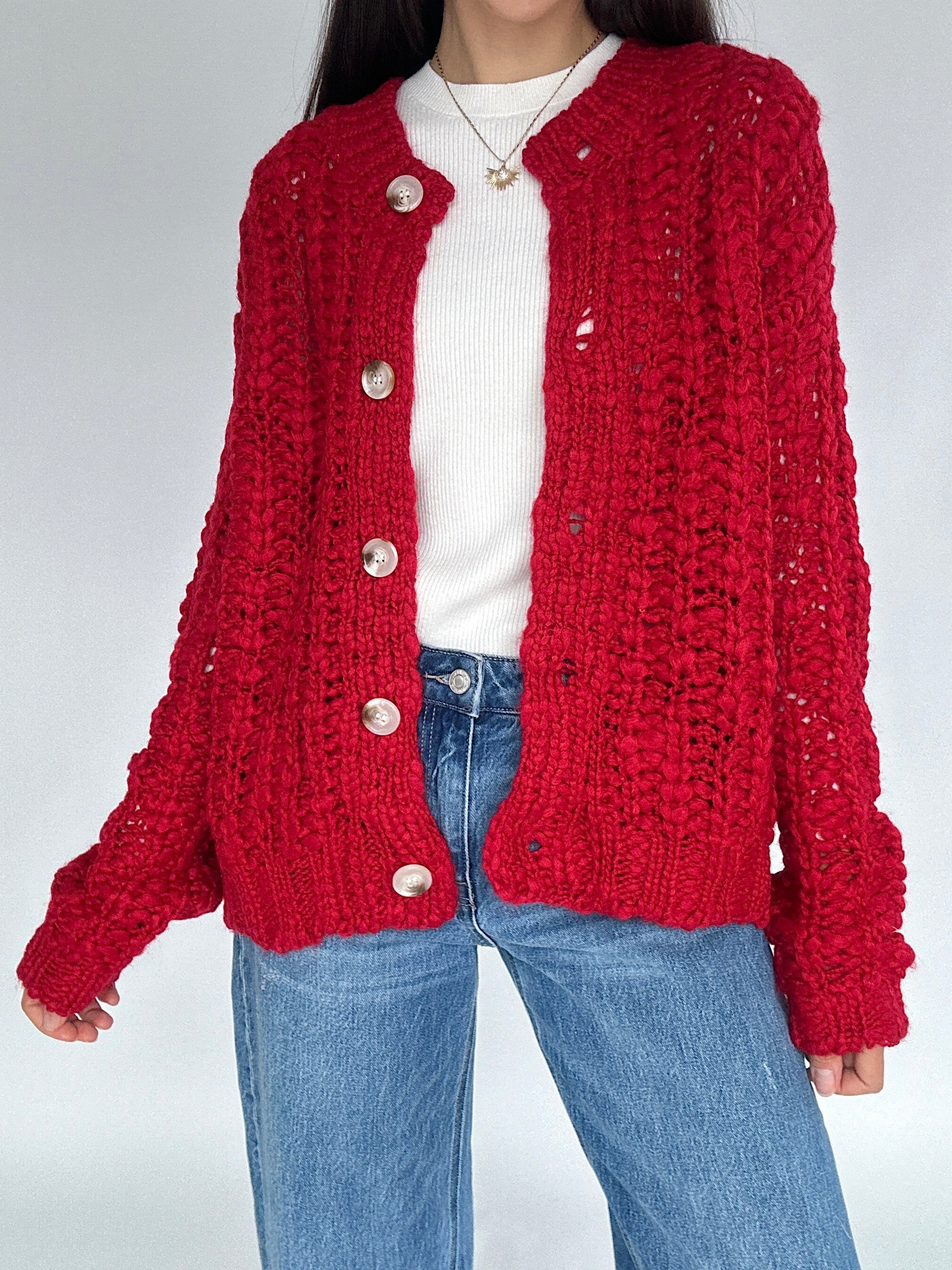 Person wearing a red knitted cardigan over a white shirt and blue jeans on a plain background