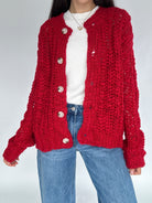 Person wearing a red knitted cardigan over a white shirt and blue jeans on a plain background