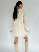 Woman wearing a white pleated dress with long sleeves on a plain background showing back