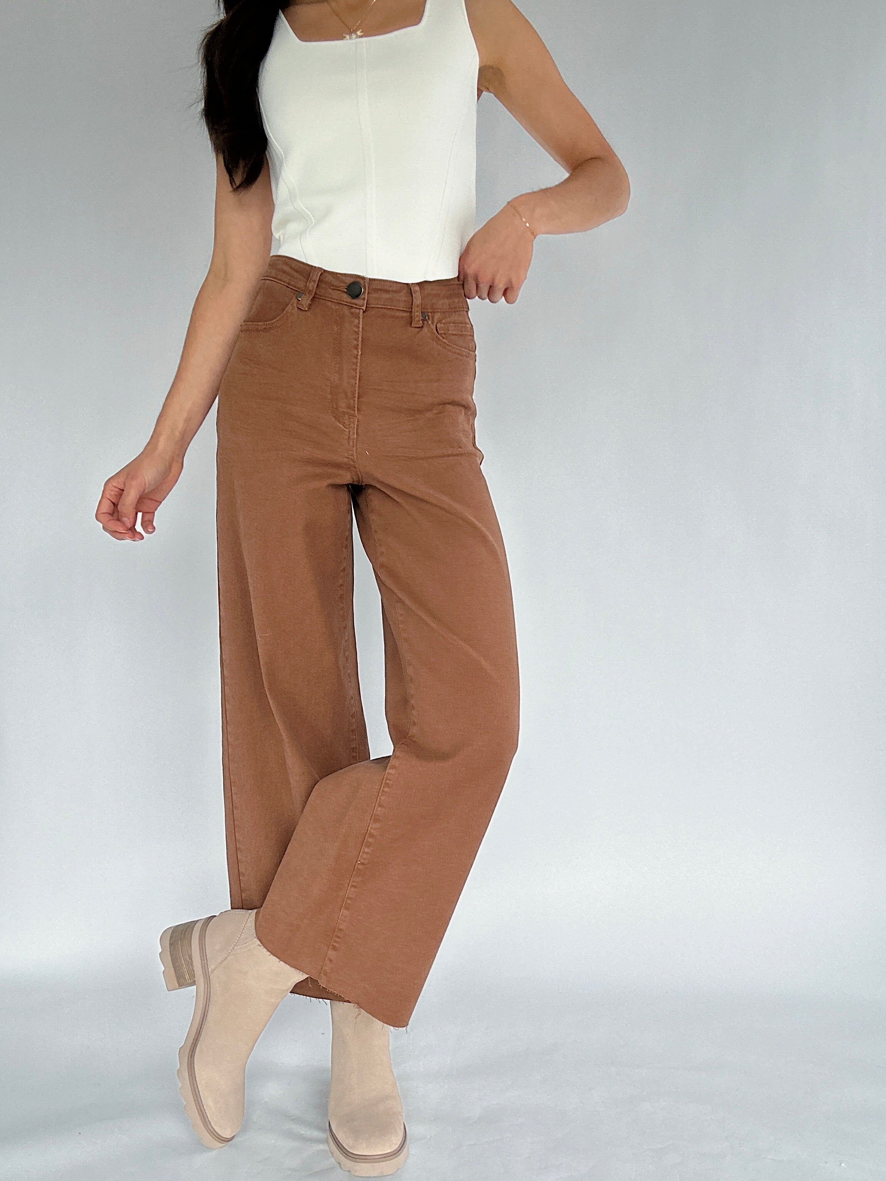 Person wearing brown wide-leg pants and a white top on a plain background with legs crossed