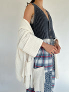 Person wearing a textured white cardigan over a navy knit vest and plaid pants with a plain background