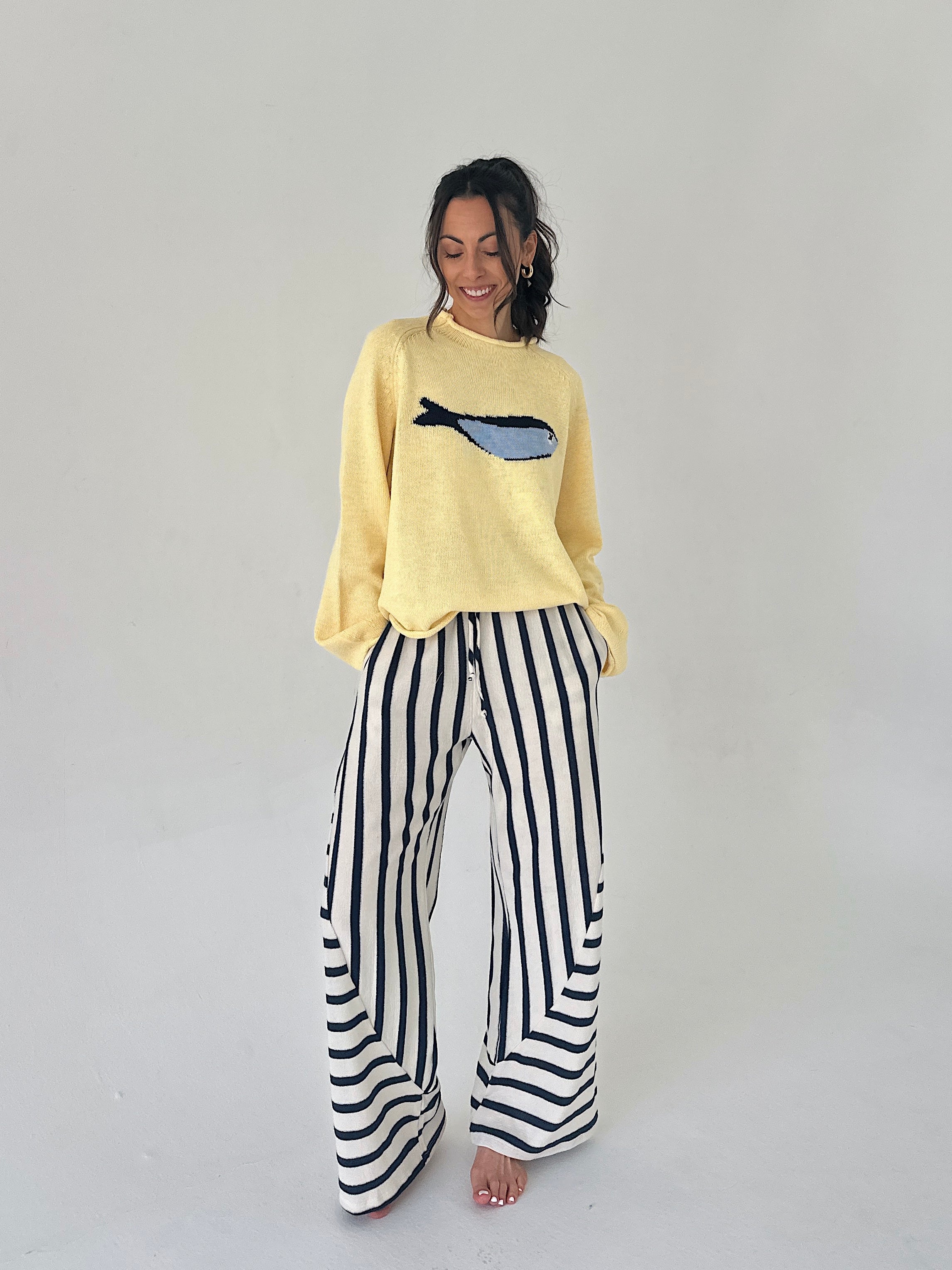 Woman wearing a yellow sweater with a fish design and black and white striped pants on a plain background
