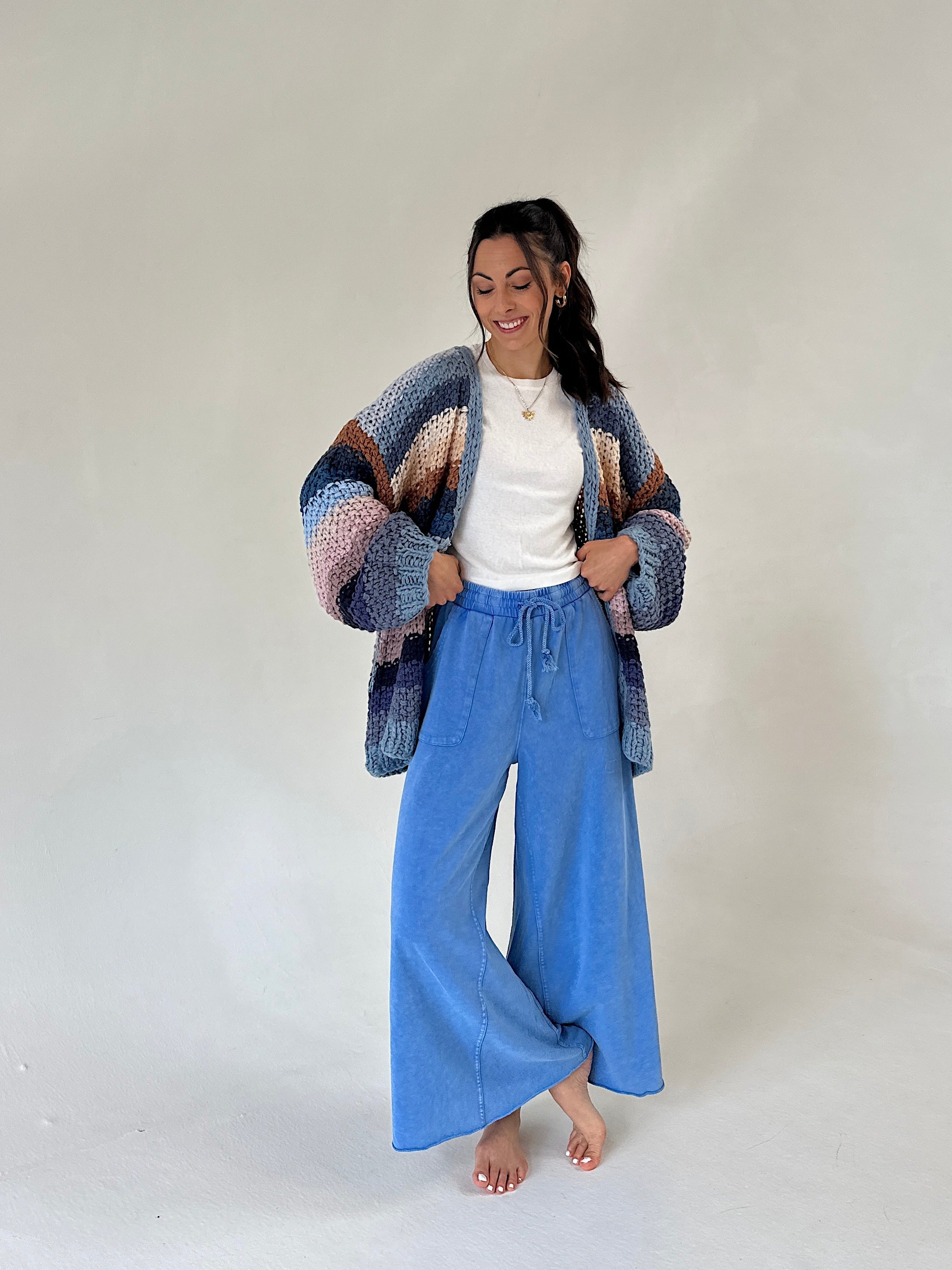 A woman stands barefoot against a plain light gray studio background, wearing a loose multicolor knit cardigan over a white top and blue wide-leg pants.