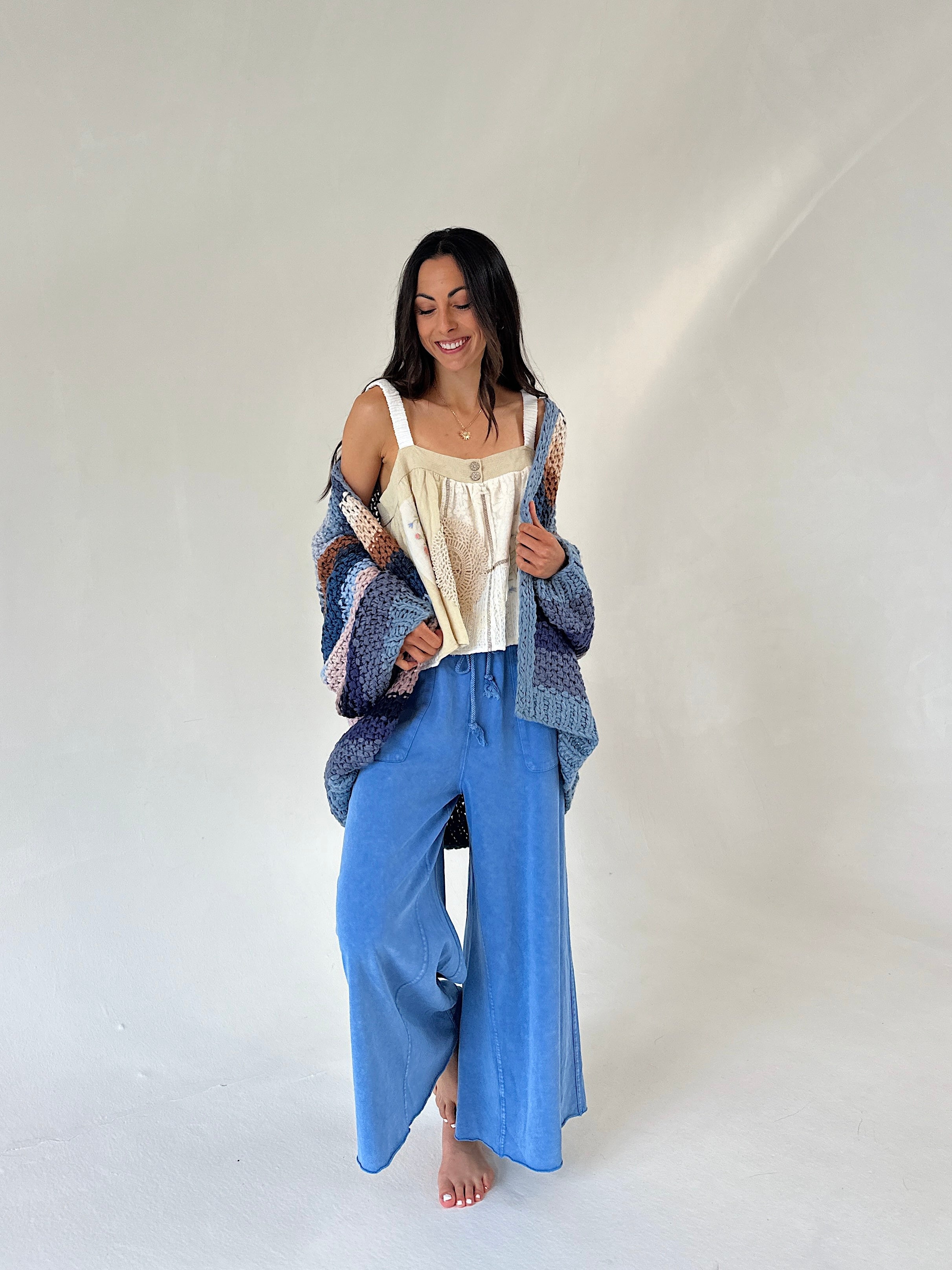 A woman stands barefoot against a plain light studio background, wearing a cream camisole top, wide-leg blue pants, and a loose patterned blue cardigan draped off her shoulders.