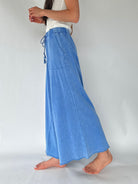 A person wearing a wide-leg pants with a drawstring waist and side pocket, paired with a white sleeveless top, standing barefoot against a plain light gray studio background.