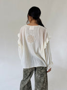 A woman viewed from behind wearing a loose white blouse with ruffled sleeves and a floral lace applique on the back, paired with gray paisley pants, against a plain light gray studio background.